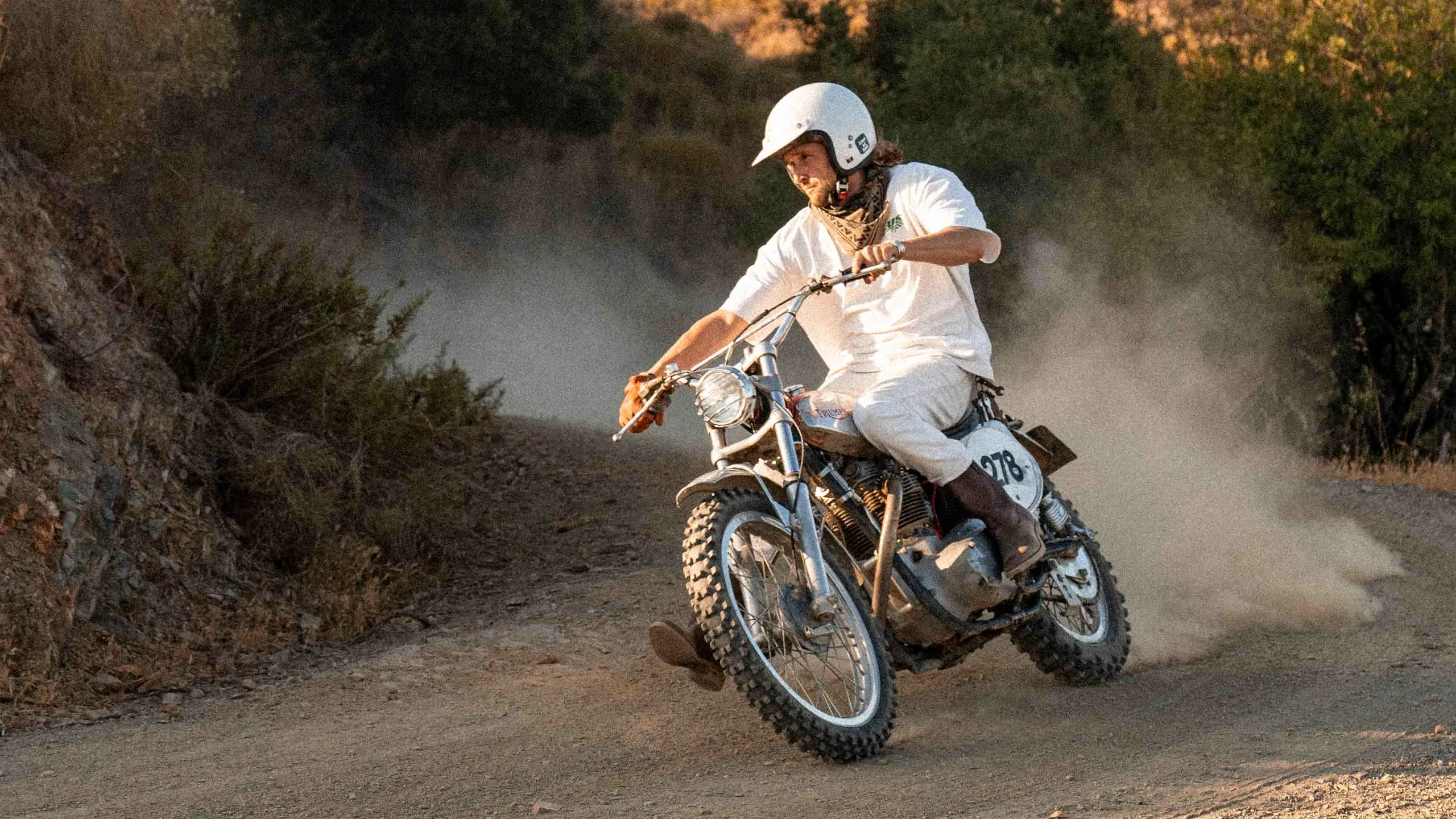 A rider powersliding a vintage motorcycle through the dust on a hill road above Mijas