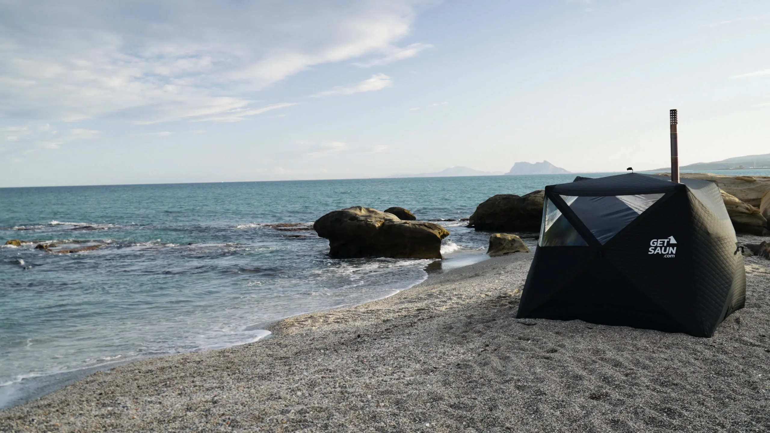 A black GetSaun sauna tent on a pebble beach, with the sea and distant mountains behind